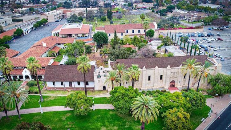 San Gabriel Mission courtyard with stone walls, arches, and lush greenery under a blue sky.